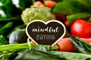 closeup of a heart-shaped chalkboard with the text mindful eating placed on a pile of some different raw vegetables, such as cucumbers, tomatoes, carrots, artichokes, green peppers and zucchinis